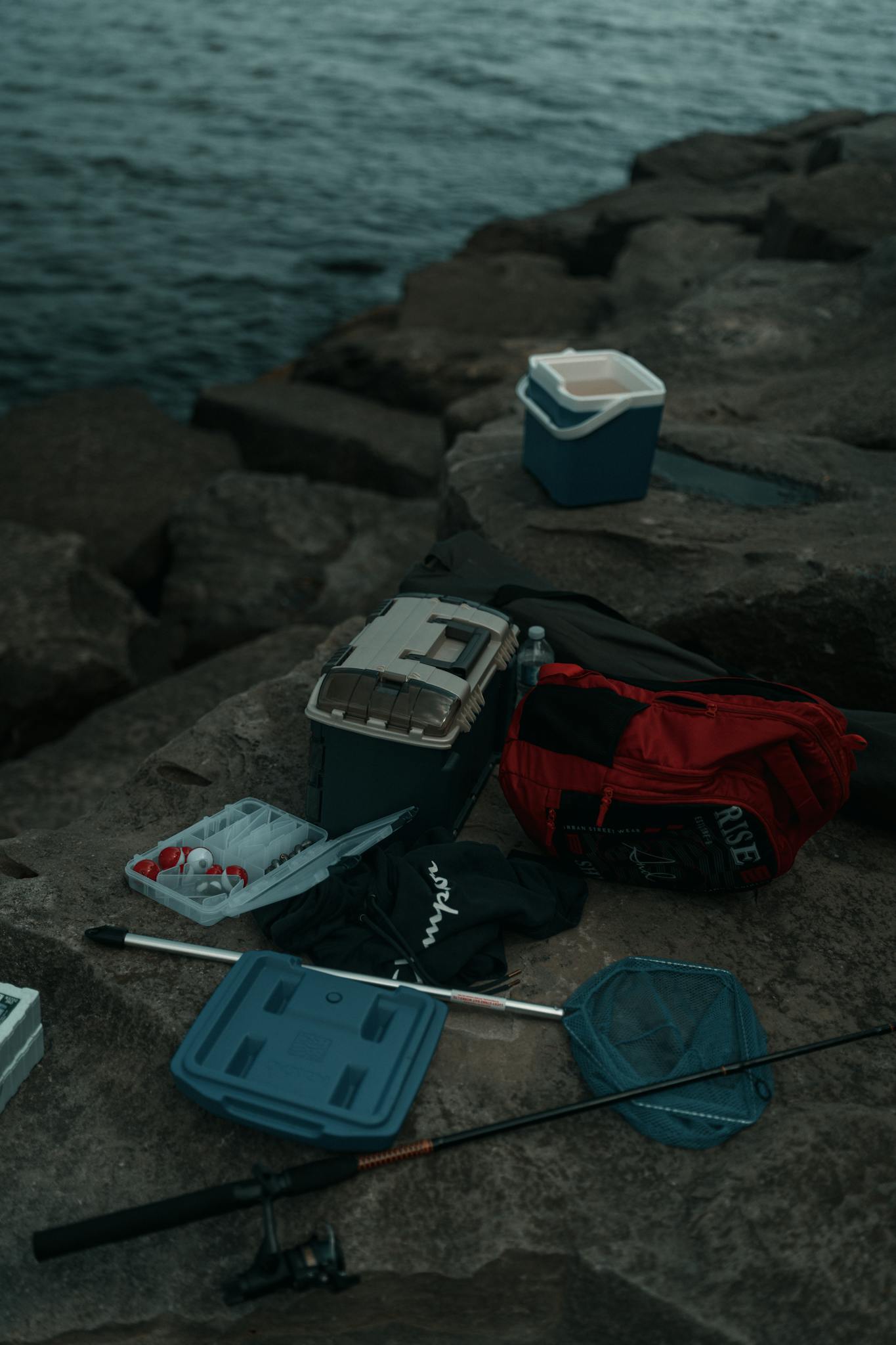 Fishing gear and supplies scattered on a rocky lakeshore during a calm evening.