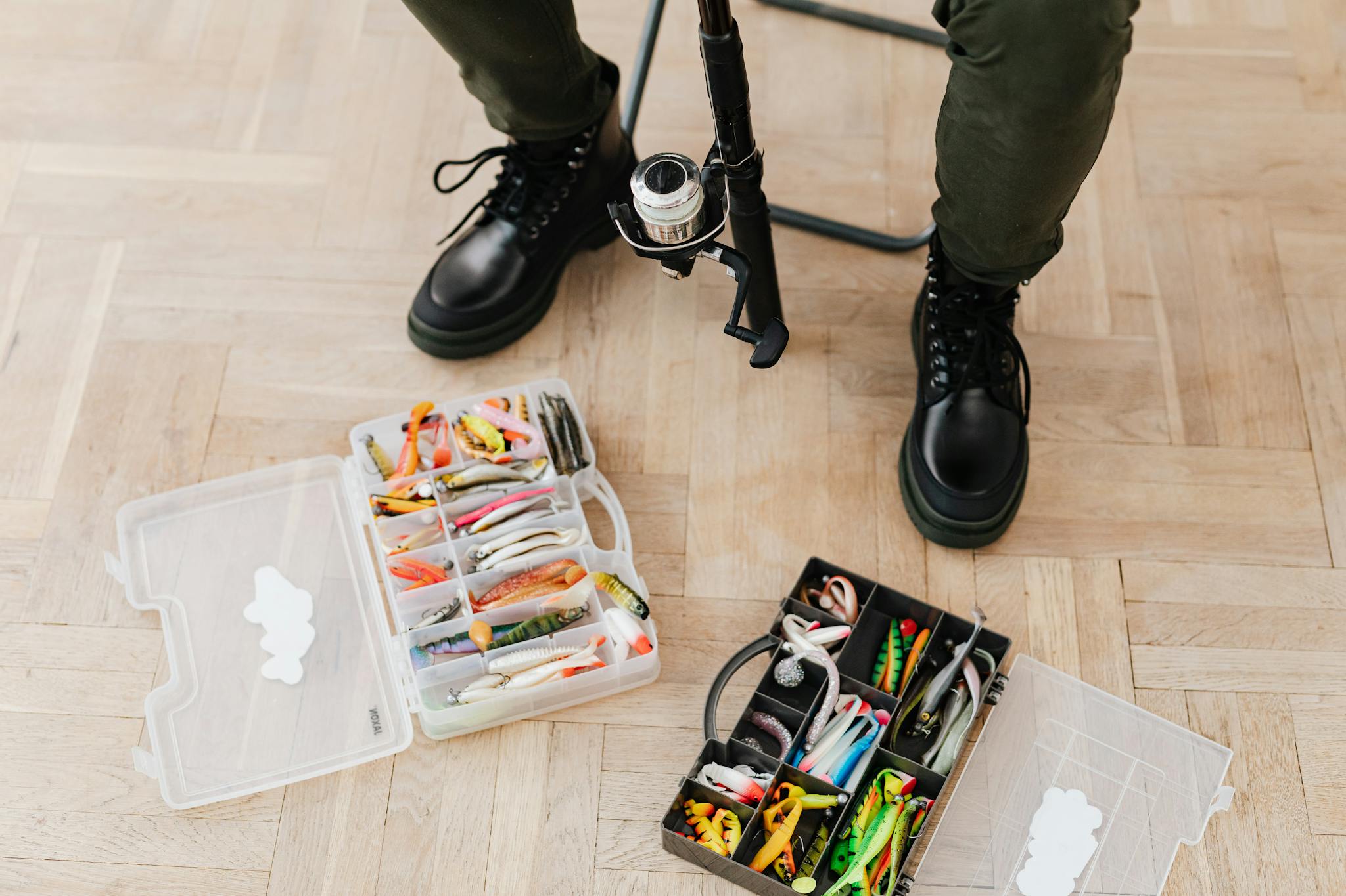 Fishing tackle boxes with lures and equipment beside boots on a wooden floor.